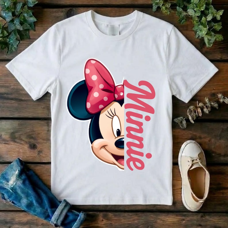 May include: A white short-sleeved t-shirt featuring a graphic of the left half of Minnie Mouse's face, complete with a pink polka-dot bow. To the right of the character graphic, the name Minnie is printed vertically in a bold pink script font.