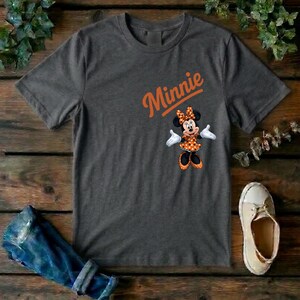 Puede incluir: Una camiseta de manga corta de color gris oscuro con el texto naranja Minnie escrito en letra cursiva sobre un gr&aacute;fico del personaje Minnie Mouse que lleva un vestido de lunares naranja y blanco y un lazo a juego sobre un fondo gris oscuro.