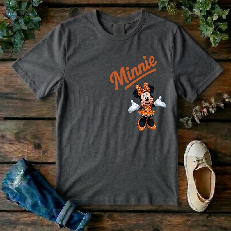 Puede incluir: Una camiseta de manga corta de color gris oscuro con el texto naranja Minnie escrito en letra cursiva sobre un gr&aacute;fico del personaje Minnie Mouse que lleva un vestido de lunares naranja y blanco y un lazo a juego sobre un fondo gris oscuro.