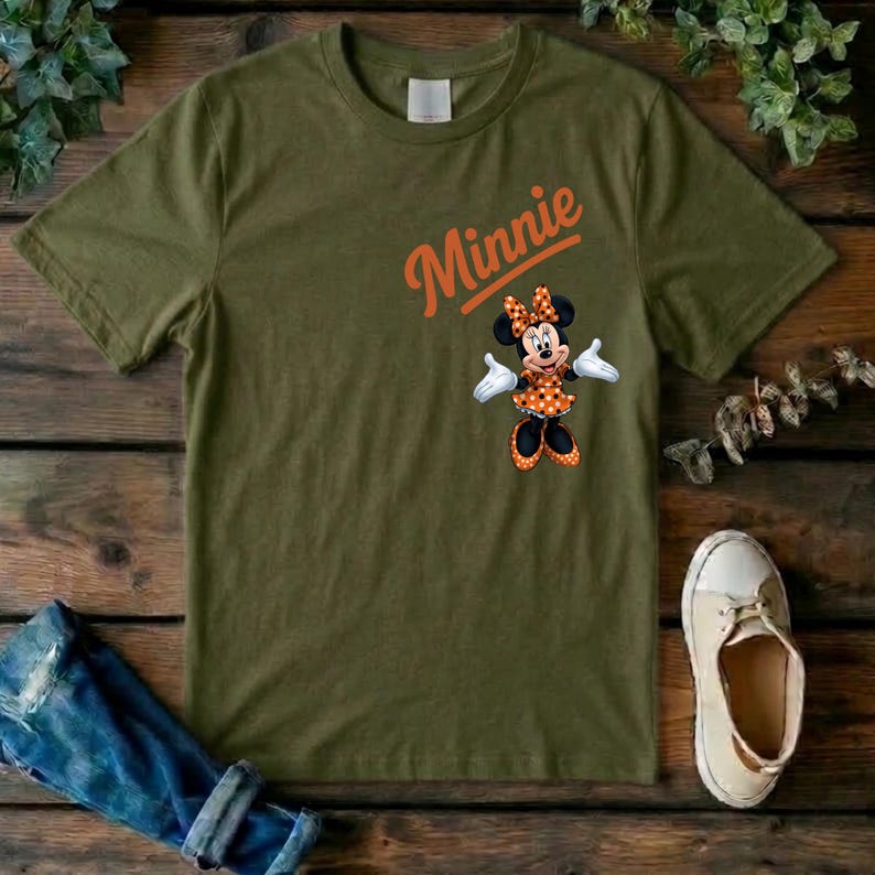 Puede incluir: Una camiseta de manga corta color verde oliva con el nombre Minnie en letras naranjas sobre un gr&aacute;fico del personaje Minnie Mouse que lleva un vestido y un lazo de lunares naranjas y blancos con guantes blancos y zapatos negros.