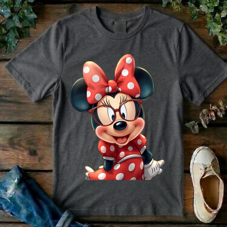 May include: A charcoal gray short-sleeve t-shirt featuring a graphic of Minnie Mouse wearing red-rimmed glasses and a red dress with white polka dots. The character has a large red bow with white polka dots on her head and white gloves.
