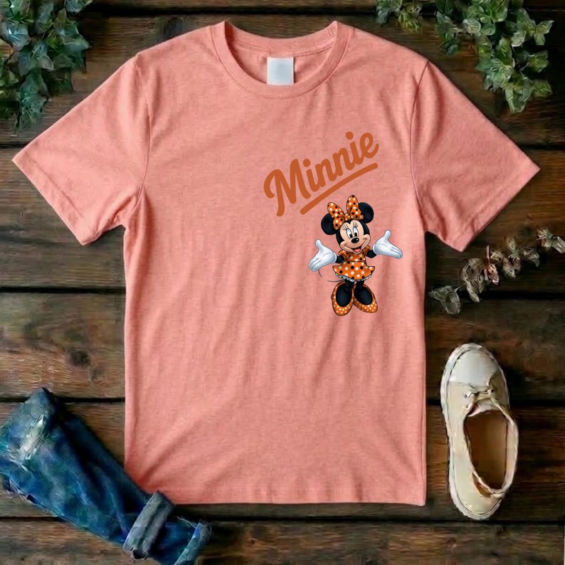 Puede incluir: Una camiseta de manga corta color melocot&oacute;n con un gr&aacute;fico de Minnie Mouse que lleva un vestido naranja de lunares y un lazo a juego. El nombre Minnie est&aacute; impreso en una fuente cursiva naranja sobre el personaje en el lado derecho del pecho.