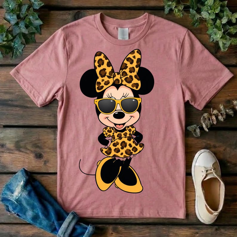 May include: A dusty rose t-shirt featuring a cartoon character wearing sunglasses, a bow, and a dress, all with a leopard print design. The character has black ears and is wearing yellow sunglasses. A pair of jeans and a shoe are visible.