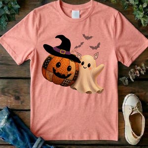 May include: A peach-colored short-sleeve t-shirt featuring a graphic print of a patchwork-style jack-o'-lantern wearing a black witch hat with a purple band, positioned next to a small, smiling cream-colored ghost with three small bats flying above.