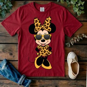 May include: Maroon t-shirt featuring a cartoon Minnie Mouse character wearing sunglasses, a bow, and a dress, all with a leopard print design. The image includes a pair of white sneakers and a pair of rolled-up blue jeans.