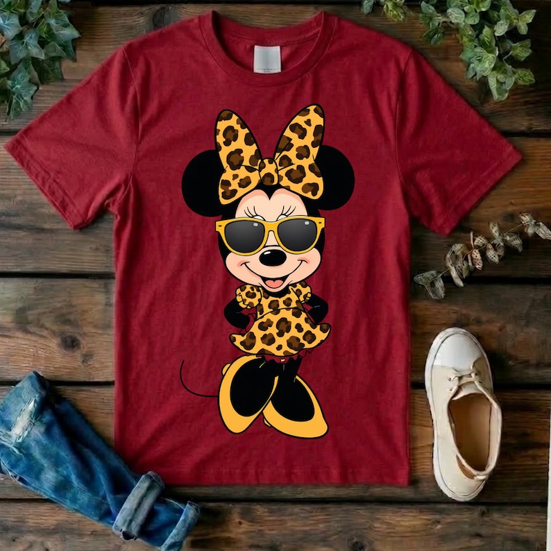 May include: Maroon t-shirt featuring a cartoon Minnie Mouse character wearing sunglasses, a bow, and a dress, all with a leopard print design. The image includes a pair of white sneakers and a pair of rolled-up blue jeans.