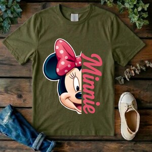 May include: An olive green short-sleeved t-shirt featuring a graphic of the right half of a cartoon mouse face with a pink polka-dot bow and the name Minnie printed vertically in pink script lettering next to the character illustration.