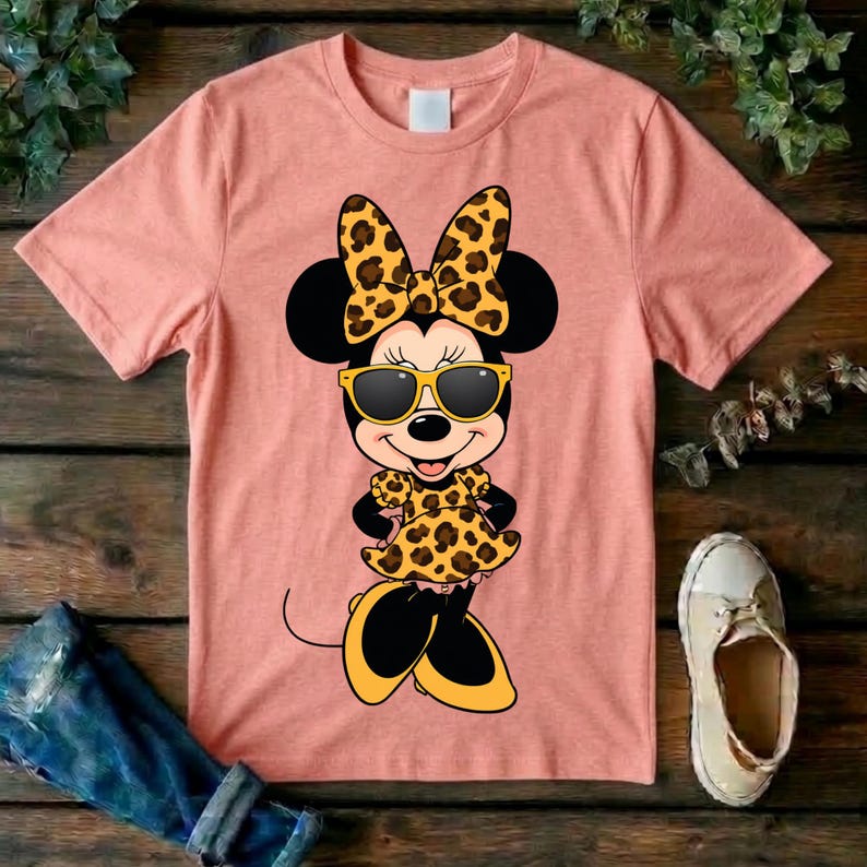 May include: Peach-colored t-shirt featuring a cartoon character with black ears, a leopard print bow, sunglasses, and a matching dress. The character's shoes are yellow and black. The shirt is on a wooden surface.