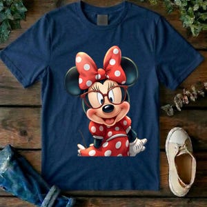 May include: A navy blue short-sleeved t-shirt featuring a graphic print of a cartoon mouse character wearing large red-rimmed glasses and a red dress with white polka dots, complemented by a matching red bow with white polka dots on the head.