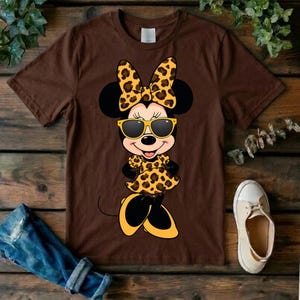 May include: Brown t-shirt featuring a cartoon Minnie Mouse character in a leopard print bow, dress, and shoes. Minnie is wearing yellow sunglasses. The shirt is on a wooden surface with a pair of jeans and a shoe.