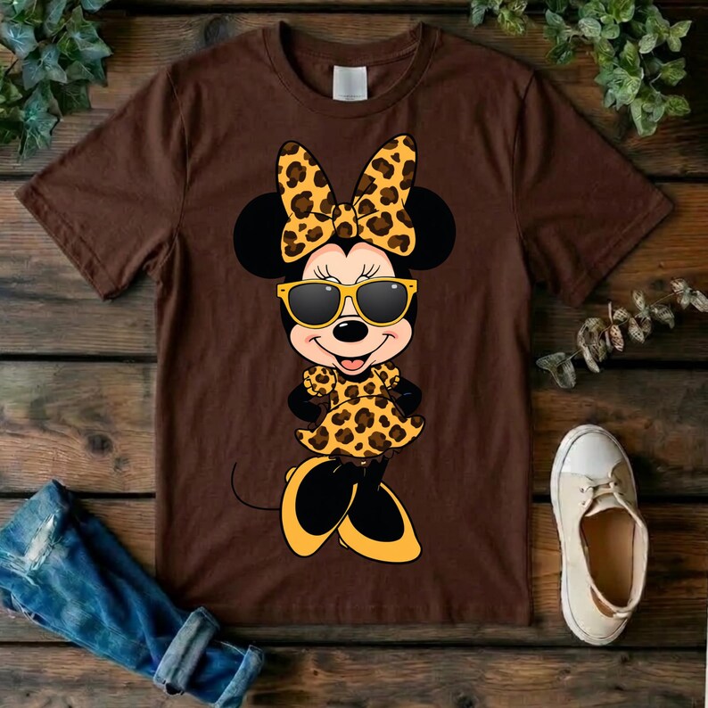 May include: Brown t-shirt featuring a cartoon Minnie Mouse character in a leopard print bow, dress, and shoes. Minnie is wearing yellow sunglasses. The shirt is on a wooden surface with a pair of jeans and a shoe.