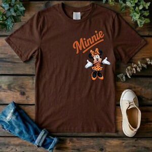 Puede incluir: Una camiseta marr&oacute;n de manga corta con un gr&aacute;fico de Minnie Mouse vistiendo un vestido naranja de lunares y un lazo. La palabra Minnie est&aacute; impresa en letra cursiva naranja sobre el personaje. La camiseta tiene cuello redondo y corte est&aacute;ndar.