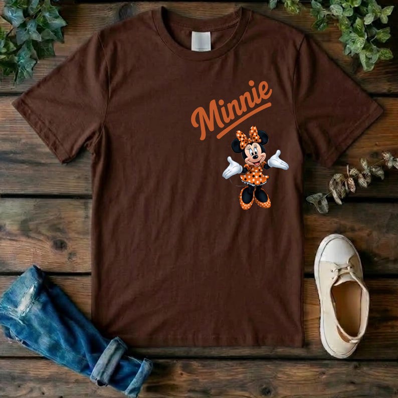 Puede incluir: Una camiseta marr&oacute;n de manga corta con un gr&aacute;fico de Minnie Mouse vistiendo un vestido naranja de lunares y un lazo. La palabra Minnie est&aacute; impresa en letra cursiva naranja sobre el personaje. La camiseta tiene cuello redondo y corte est&aacute;ndar.