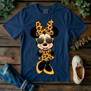 May include: Navy blue t-shirt featuring a cartoon Minnie Mouse character wearing sunglasses and a leopard print bow, dress, and shoes. The graphic is centered on the front of the shirt. The shirt is made of a soft material.