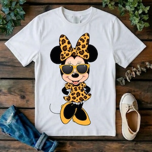 May include: White t-shirt featuring a cartoon Minnie Mouse character wearing sunglasses, a bow, and a dress, all with a leopard print design. The character has black ears and is smiling. The shirt is on a wooden surface.