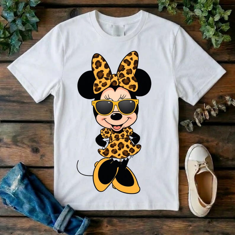 May include: White t-shirt featuring a cartoon Minnie Mouse character wearing sunglasses, a bow, and a dress, all with a leopard print design. The character has black ears and is smiling. The shirt is on a wooden surface.