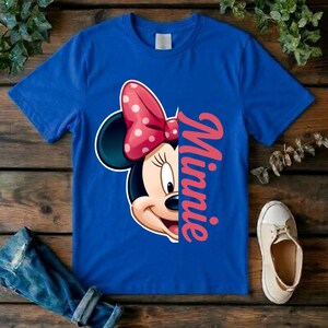 May include: A royal blue short-sleeved t-shirt featuring a graphic of half of Minnie Mouse's face with a red and white polka dot bow. The name Minnie is printed in a vertical, cursive pink font next to the character illustration on the front.