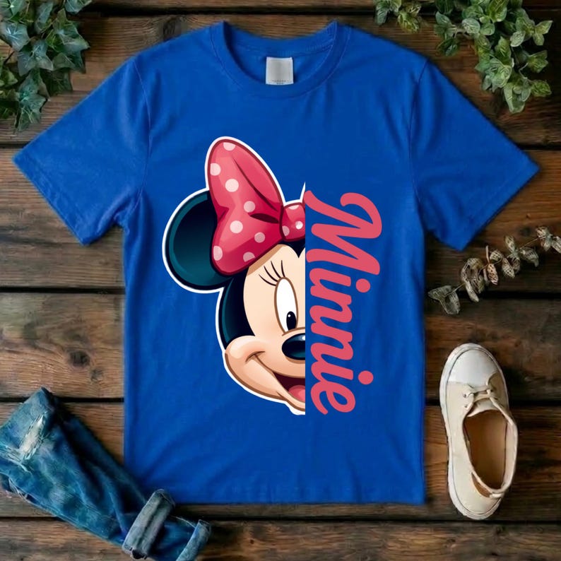 May include: A royal blue short-sleeved t-shirt featuring a graphic of half of Minnie Mouse's face with a red and white polka dot bow. The name Minnie is printed in a vertical, cursive pink font next to the character illustration on the front.