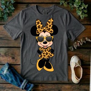 May include: A charcoal gray t-shirt featuring a cartoon Minnie Mouse character in a leopard print bow, dress, and sunglasses. The image is set against a wooden background with a pair of jeans and a shoe.