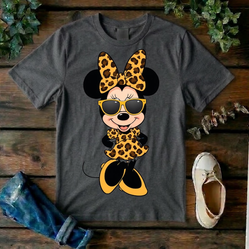May include: A charcoal gray t-shirt featuring a cartoon Minnie Mouse character in a leopard print bow, dress, and sunglasses. The image is set against a wooden background with a pair of jeans and a shoe.
