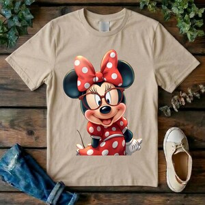 May include: A beige short-sleeved t-shirt featuring a graphic print of a cartoon mouse character wearing red-rimmed glasses and a red dress with white polka dots, topped with a matching red bow with white polka dots on its head.