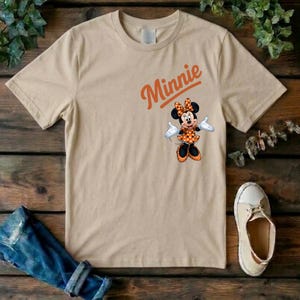 Puede incluir: Una camiseta beige de manga corta con la palabra Minnie en texto naranja sobre un gr&aacute;fico del personaje Minnie Mouse con un vestido y un lazo de lunares naranjas y blancos, guantes blancos y zapatos negros.