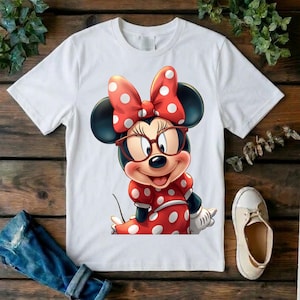 May include: A white short-sleeved t-shirt featuring a graphic print of a cartoon mouse character wearing red-rimmed glasses and a red dress with white polka dots, complemented by a matching red bow with white polka dots on the head.