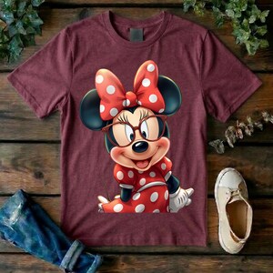 May include: A maroon short-sleeved t-shirt featuring a graphic of Minnie Mouse wearing large brown-rimmed glasses and a red bow with white polka dots. The character is also wearing a matching red dress with white polka dots and white gloves.