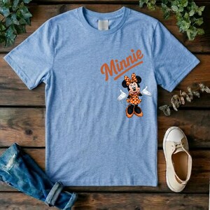 Puede incluir: Una camiseta de color azul claro jaspeado con el nombre Minnie impreso en letra cursiva naranja sobre un gr&aacute;fico del personaje Minnie Mouse, que lleva un vestido y un lazo de lunares naranjas y blancos, con guantes blancos y zapatos negros.