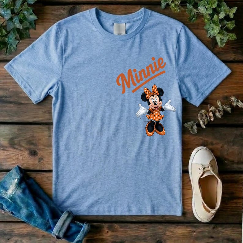 Puede incluir: Una camiseta de color azul claro jaspeado con el nombre Minnie impreso en letra cursiva naranja sobre un gr&aacute;fico del personaje Minnie Mouse, que lleva un vestido y un lazo de lunares naranjas y blancos, con guantes blancos y zapatos negros.