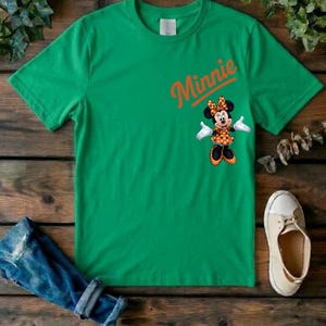 Puede incluir: Una camiseta verde brillante de manga corta con el nombre Minnie escrito en letra cursiva naranja sobre un gr&aacute;fico del personaje Minnie Mouse con un vestido de lunares naranja y blanco y un lazo a juego sobre un fondo liso.