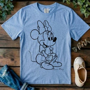 May include: A light blue short-sleeve t-shirt featuring a black line art graphic of Minnie Mouse sitting down. The character is depicted with her signature bow and dress. The design is centered on the chest of the garment, which has a crew neckline.