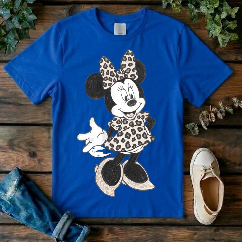 Op de afbeelding: Een koningsblauw T-shirt met korte mouwen en een afbeelding van een tekenfilmmuis. Het personage draagt een jurk en een strik, beide versierd met een bruin en zwart luipaardpatroon. De muis heeft zwarte oren en witte handschoenen.