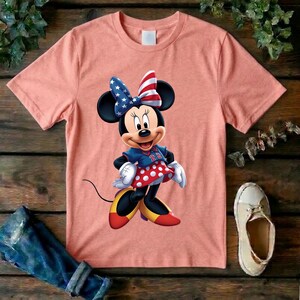 May include: A peach-colored short-sleeve t-shirt featuring a graphic of Minnie Mouse wearing a blue jersey with the number 22, a red and white polka dot skirt, and a bow patterned with stars and stripes. The character wears yellow shoes.