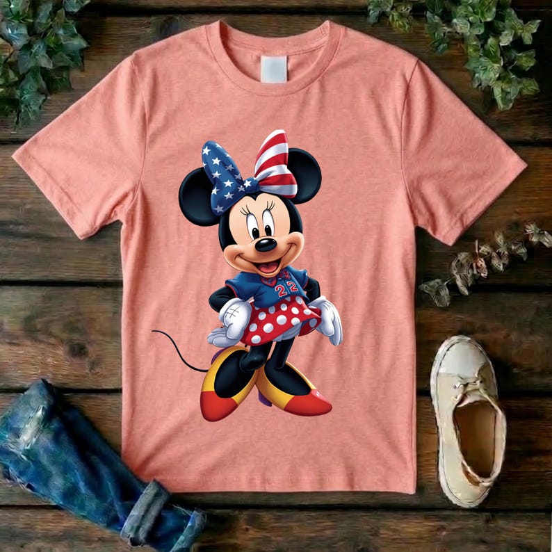 May include: A peach-colored short-sleeve t-shirt featuring a graphic of Minnie Mouse wearing a blue jersey with the number 22, a red and white polka dot skirt, and a bow patterned with stars and stripes. The character wears yellow shoes.