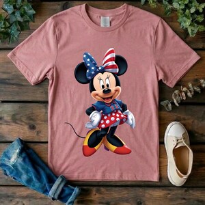 May include: A dusty rose short-sleeved t-shirt featuring a graphic of a cartoon mouse wearing a blue and red star-patterned bow, a blue jersey with the number 22, and a red skirt with white polka dots, paired with yellow and red shoes.