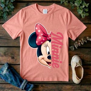 May include: A peach-colored short-sleeve t-shirt featuring a graphic of half of Minnie Mouse's face with a pink polka-dot bow. The text Minnie is printed vertically in a pink script font next to the character graphic on the front of the shirt.