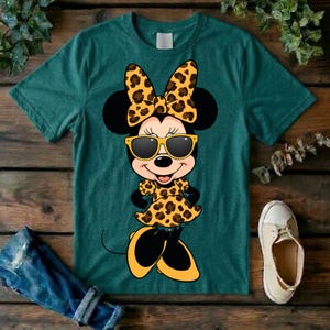 May include: A teal t-shirt featuring a cartoon character with black ears, a bow, sunglasses, and a dress, all with a leopard print design. The character's shoes are yellow and black. A pair of blue jeans and a white shoe are visible.