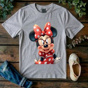 May include: A heather gray short-sleeve t-shirt featuring a graphic print of a cartoon mouse character wearing red-rimmed glasses and a red dress with white polka dots, complemented by a matching red bow with white polka dots on the head.