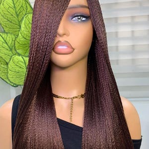 May include: A brown braided wig with long, straight strands. The wig is styled with a side part and frames the face. The mannequin is wearing a black top and a gold necklace.
