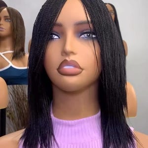 May include: A mannequin head with a black braided wig. The wig features a center part and falls to the shoulders. The mannequin is wearing a light purple ribbed top. Other mannequin heads with wigs are visible in the background.