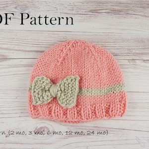 May include: PDF pattern for a knitted baby hat with a bow. The hat is pink with a beige stripe and a beige bow. The pattern includes sizes for newborn, 2 months, 3 months, 6 months, 12 months, and 24 months.