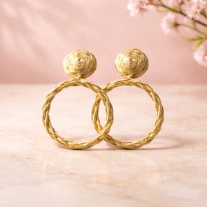 Puede incluir: Un par de pendientes de aro dorados con un diseño tejido. Cada pendiente presenta un aro circular y un pequeño detalle redondo en la parte superior. Los pendientes se muestran sobre un fondo rosa suave, ideal para joyería o moda.