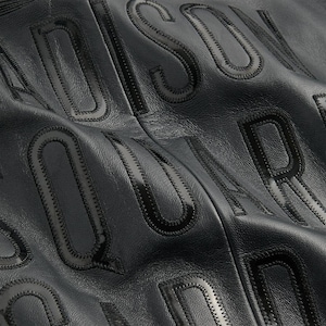 May include: Close-up of black leather material with embossed lettering. The letters are raised and have a glossy finish, with a stitched outline. The background is a matte black, creating a contrast with the glossy letters.