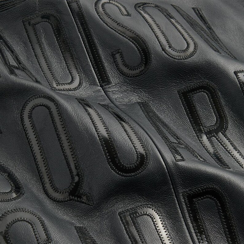 May include: Close-up of black leather material with embossed lettering. The letters are raised and have a glossy finish, with a stitched outline. The background is a matte black, creating a contrast with the glossy letters.