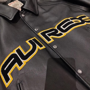 May include: Black leather jacket featuring the word "AUREX" in large, yellow-outlined, black lettering across the chest. The jacket has a collared neckline, snap closures, and a small pocket. The jacket appears to be new.