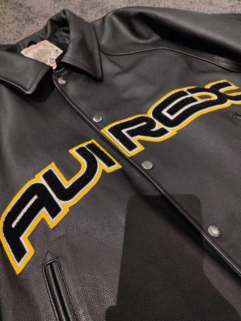 May include: Black leather jacket featuring the word "AUREX" in large, yellow-outlined, black lettering across the chest. The jacket has a collared neckline, snap closures, and a small pocket. The jacket appears to be new.