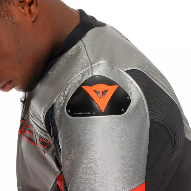May include: Close-up of a silver and black motorcycle jacket with red accents. The jacket features a black patch with an orange logo and the text "ENGINEERED TO SLIDE". The jacket is made of leather.