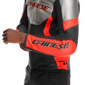 May include: A black and gray motorcycle jacket with red accents. The jacket features the brand name "DAINESE" in large letters on the sleeve and "DAINESE" on the shoulder. The jacket is made of leather and has a sporty design.