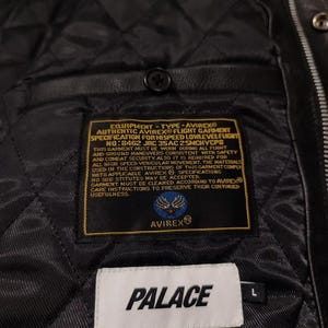 May include: Black leather jacket interior with a quilted lining. Features labels with "AVIREX" and "PALACE" branding. The jacket has a button closure and a size "L" tag.
